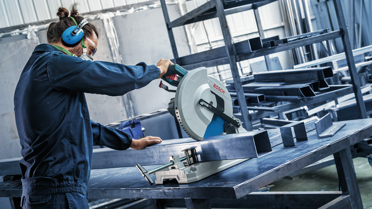 Person wearing safety equipment uses a circular saw to cut a metal beam on a worktable.