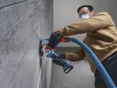 A person wearing safety equipment uses an angle grinder with dust extraction on a concrete wall.