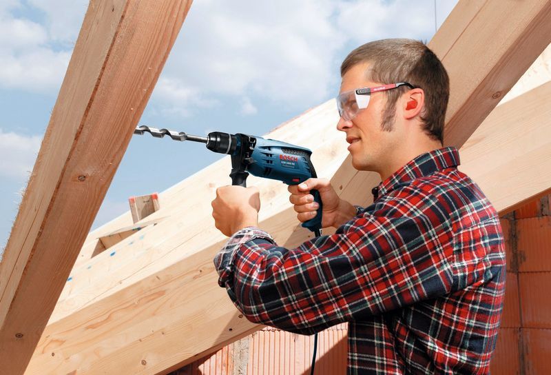 A person wearing safety equipment drills into a wooden beam at a construction site.