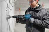A person wearing safety equipment drills a large hole in a concrete wall using a power tool.