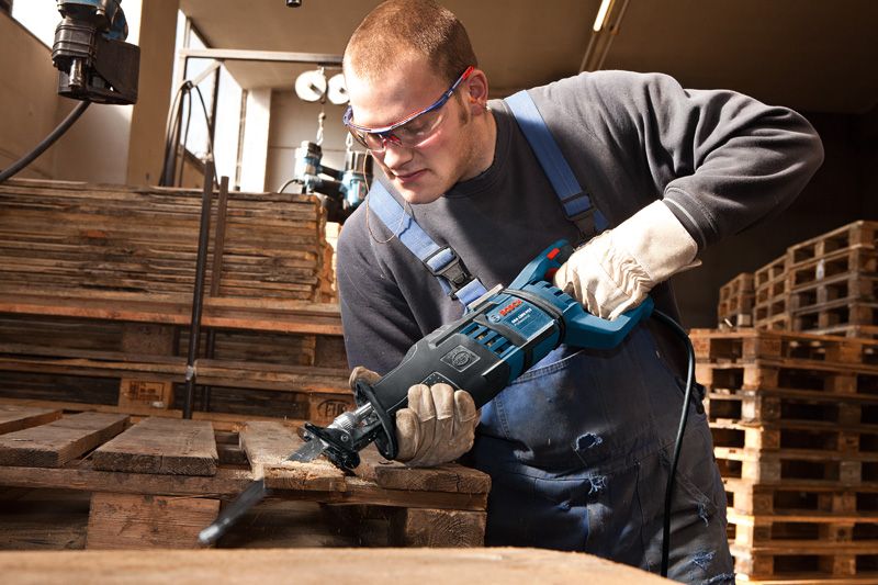 A person wearing safety equipment uses a reciprocating saw to cut a wooden pallet.