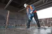 A person wearing safety equipment drills into concrete with a rotary hammer.