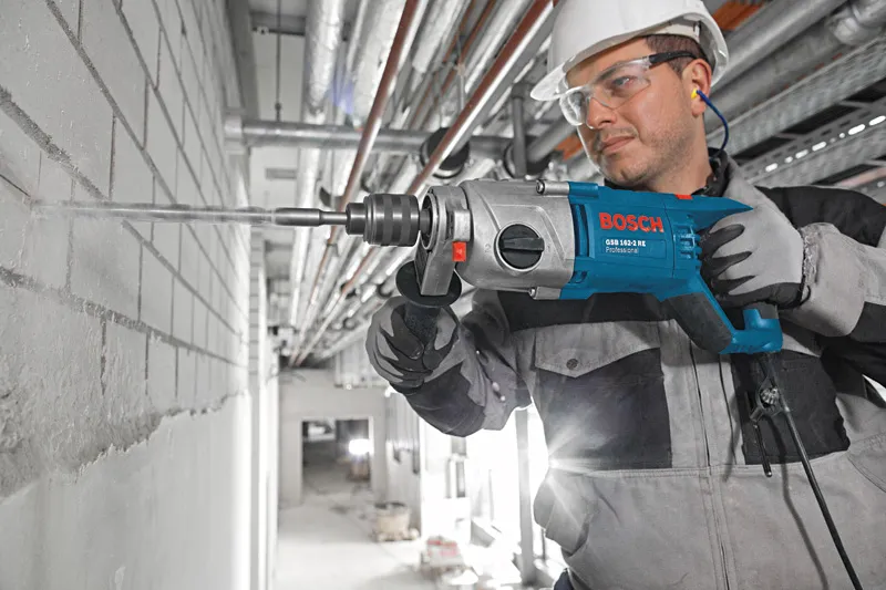 Electric impact drill with auxiliary handle and long masonry bit attached.
