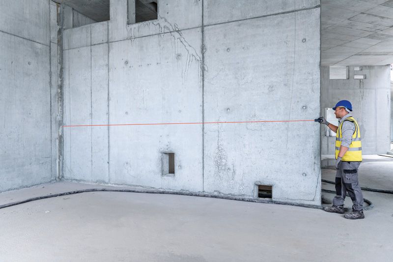 A person wearing safety equipment measures a concrete wall with a laser measure.