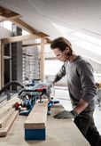 A person wearing safety equipment cuts a wooden board with a circular saw in a workshop.
