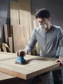 Person wearing safety equipment sands a wooden door with an orbital sander in a workshop.