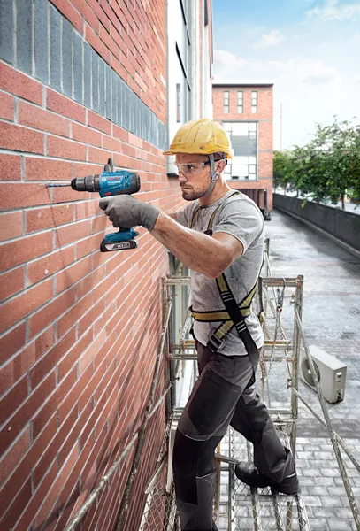 A person wearing safety equipment drills into a brick wall on scaffolding.