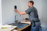 A person uses a detector on a kitchen wall above a sink.