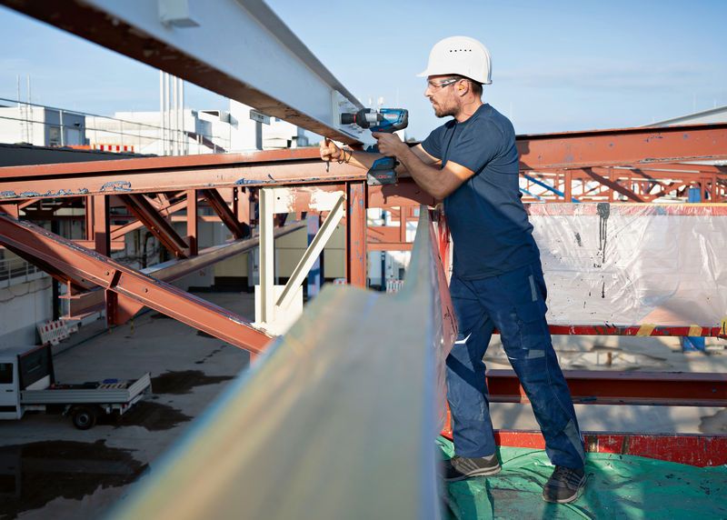 A person wearing safety equipment uses a cordless impact wrench on a steel beam.