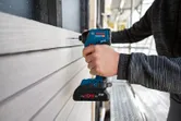A person uses a cordless power drill to fasten screws into wooden siding.