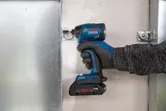 A person wearing safety equipment uses a cordless impact driver to fasten a metal bracket.