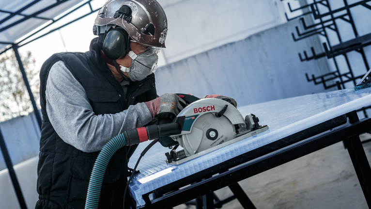 A person wearing safety equipment uses a circular saw to cut a sheet on a workbench.