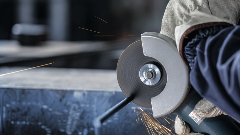 Person wearing safety equipment cuts metal with an angle grinder, creating sparks.
