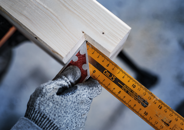 Person wearing safety equipment measures a wooden beam with a square ruler.