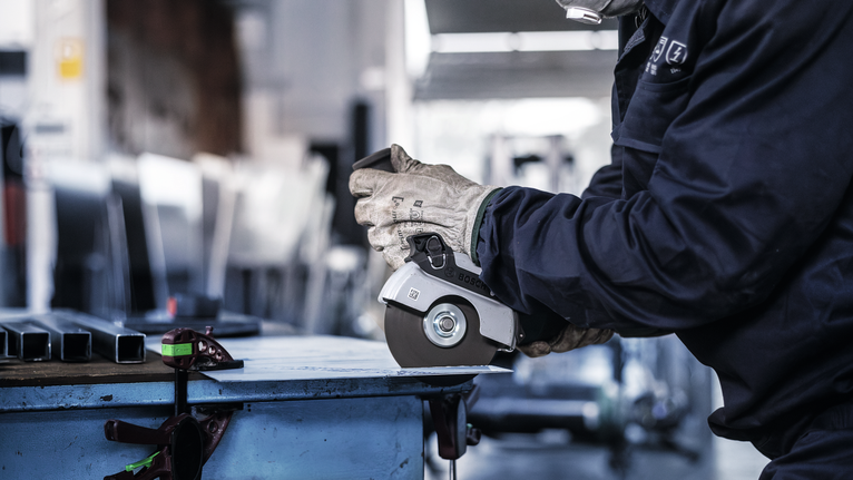 A person wearing safety equipment uses an angle grinder to cut metal.