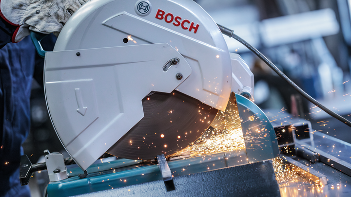 A person wearing safety equipment cuts metal with a circular saw, producing bright sparks.