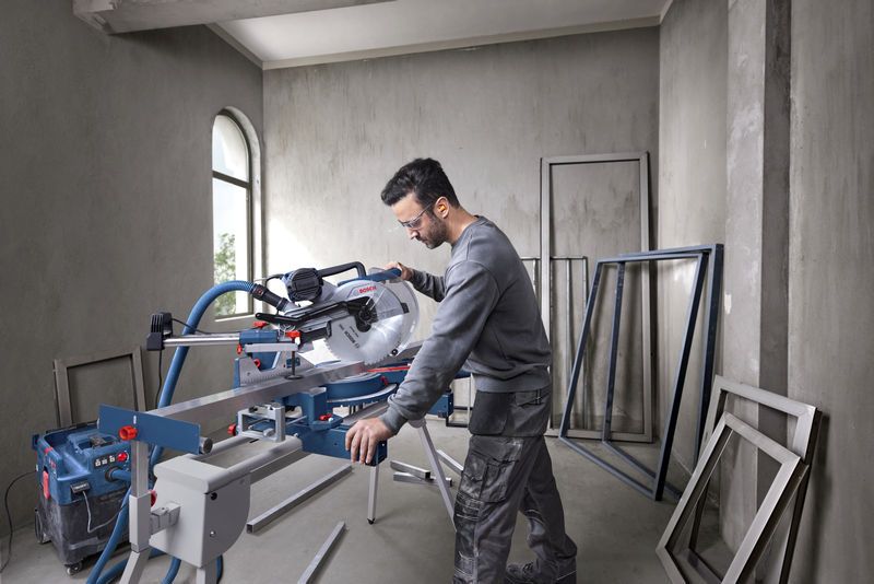 A person wearing safety equipment operates a mitre saw to cut metal frames indoors.