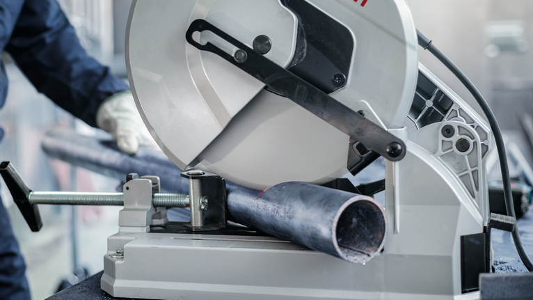 A person wearing safety equipment cuts a metal pipe with a stationary saw.