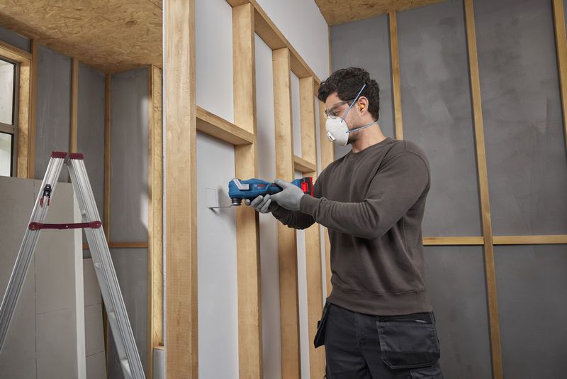 A person wearing safety equipment uses a multifunction tool to cut drywall.