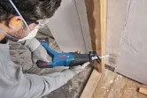 A person wearing safety equipment cuts a wooden beam with a cordless reciprocating saw.