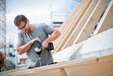 الشخص الذي يرتدي معدات السلامة يستخدم مفتاح ربط لاسلكي لدفع المسمار في الخشب.