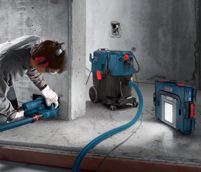 A person wearing safety equipment uses a tool connected to a corded dust extractor.