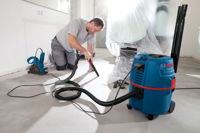 A person kneels on the floor using a corded dust extractor to clean up debris.