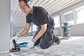 A person wearing safety equipment grinds a concrete floor with a blue concrete grinder.