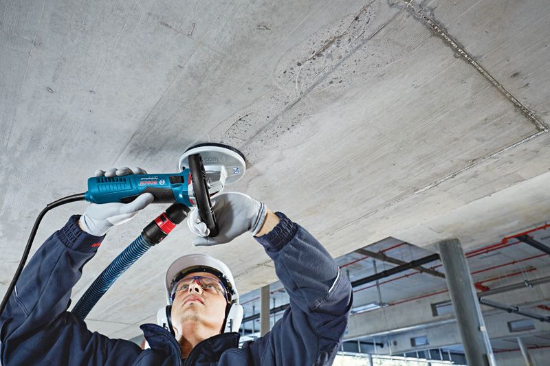 A person wearing safety equipment grinds a concrete ceiling with a concrete grinder.