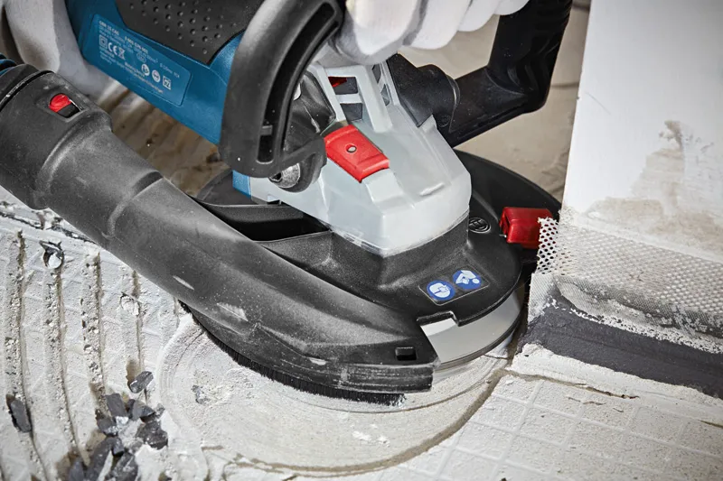 A person wearing safety equipment grinds concrete floor with a concrete grinder near a wall.
