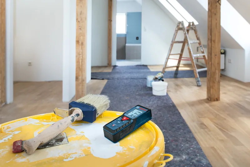 Laser measure and paintbrush on a yellow bucket in a room under renovation.