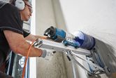 A person wearing safety equipment drills into a wall using a mounted drill stand.