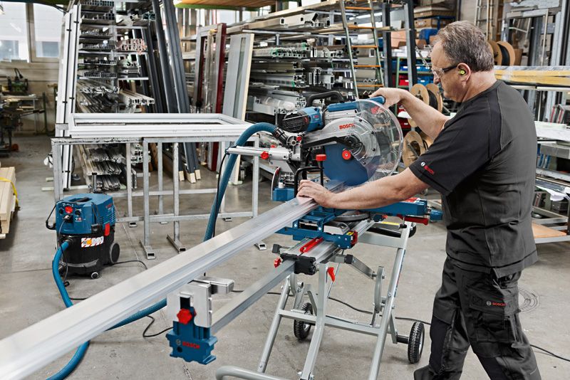 A person wearing safety equipment operates a sliding mitre saw to cut a metal profile.