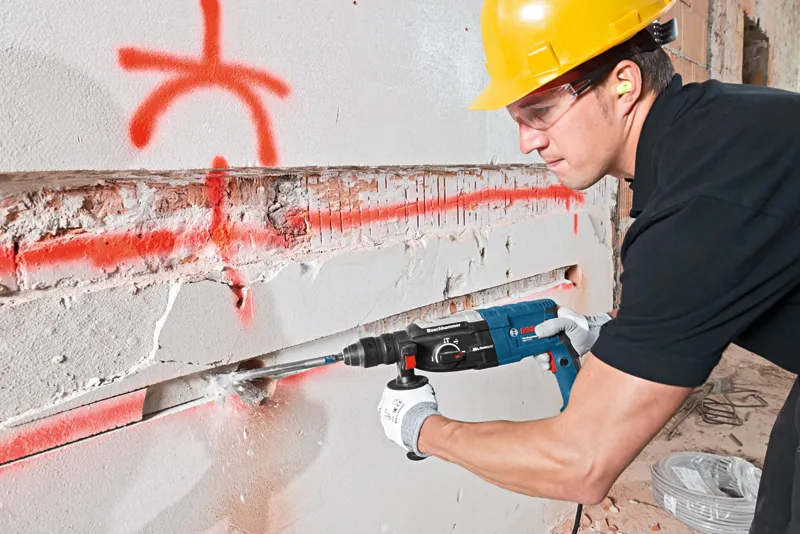 A person wearing safety equipment uses a rotary hammer to chisel a groove in a concrete wall.