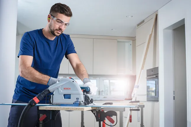 Person wearing safety equipment cuts wood panel with a track saw in a modern kitchen.