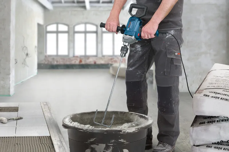 A person wearing safety equipment mixes mortar in a bucket with a power mixer.