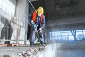 A person wearing safety equipment uses a demolition hammer to break concrete indoors.