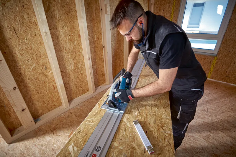 A person wearing safety equipment uses a circular saw with a guide rail to cut wood.