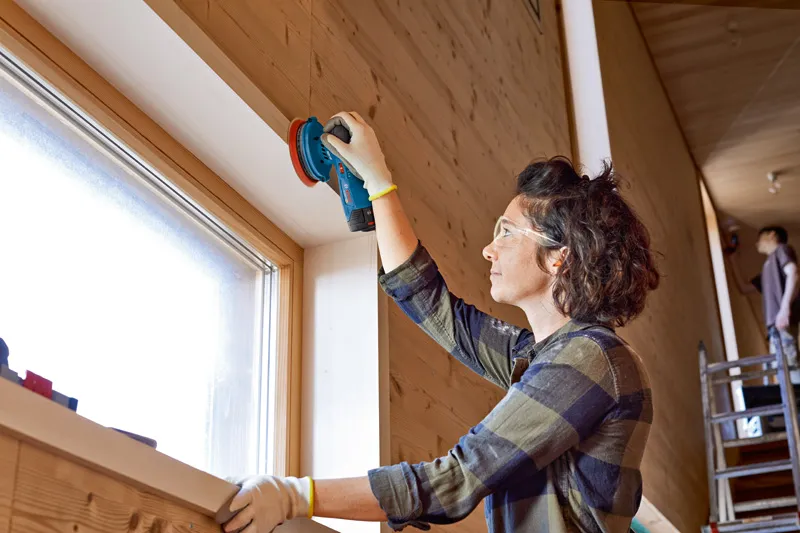 A person wearing safety equipment sands a wooden window frame with a cordless random orbit sander.