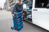 A person wearing safety equipment loads stacked toolboxes onto a dolly beside a work van.
