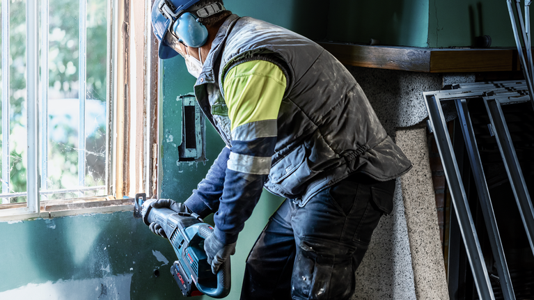 Person wearing safety equipment uses a power tool to cut a window frame.
