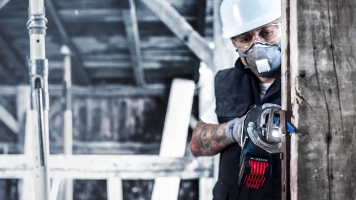 A person wearing safety equipment uses a cordless angle grinder to cut through wood.