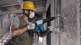 A person wearing safety equipment uses a demolition hammer to break a concrete wall.