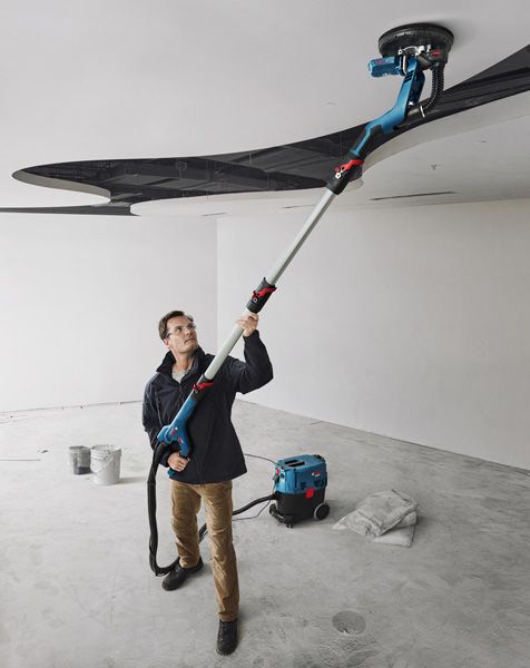 A person wearing safety equipment sands a ceiling with a drywall sander in a renovation space.
