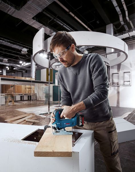 A person wearing safety equipment uses a cordless jigsaw to cut wood indoors.