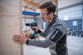 Person wearing safety equipment drills into a wooden board inside a workshop.