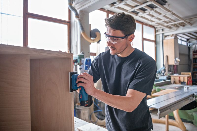 Person wearing safety equipment sands wooden furniture with a cordless sander.