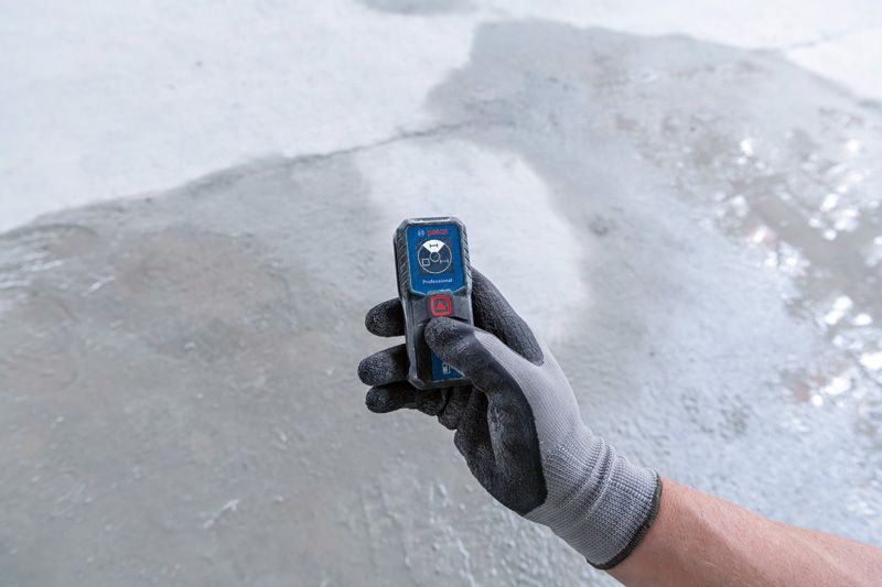 Person wearing safety equipment holds a laser measure on a concrete floor.