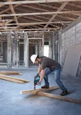 A person wearing safety equipment drills into a wooden beam at a construction site.