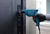 Person wearing safety equipment uses a power drill to fasten screws into a metal doorframe.